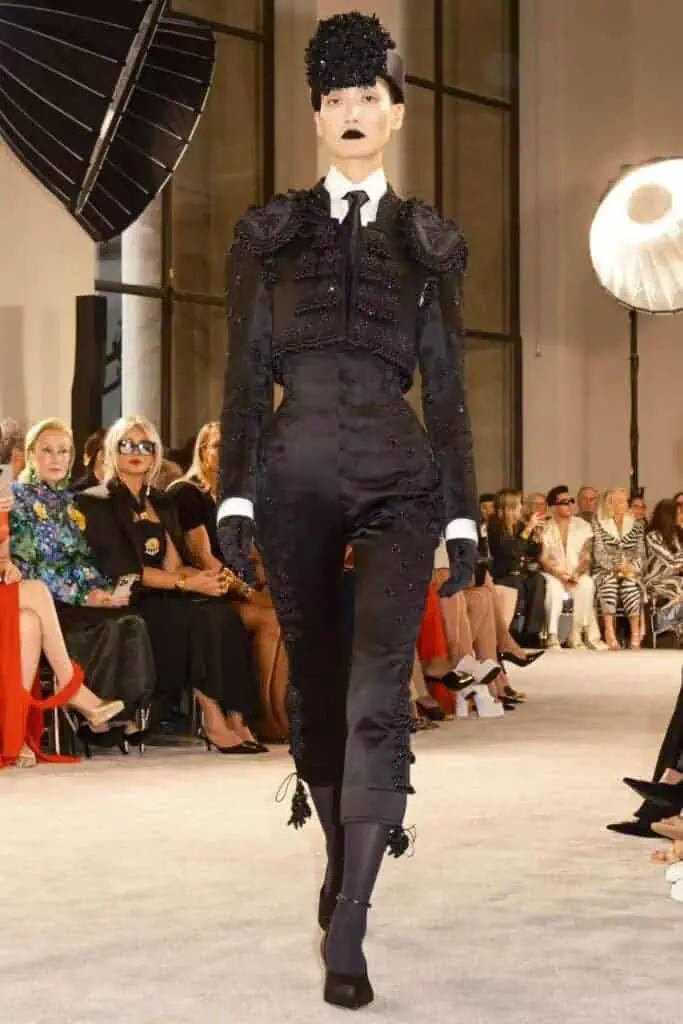 Model wearing a black embellished matador-style suit with exaggerated hip structure, black gloves, and dramatic headpiece on the runway