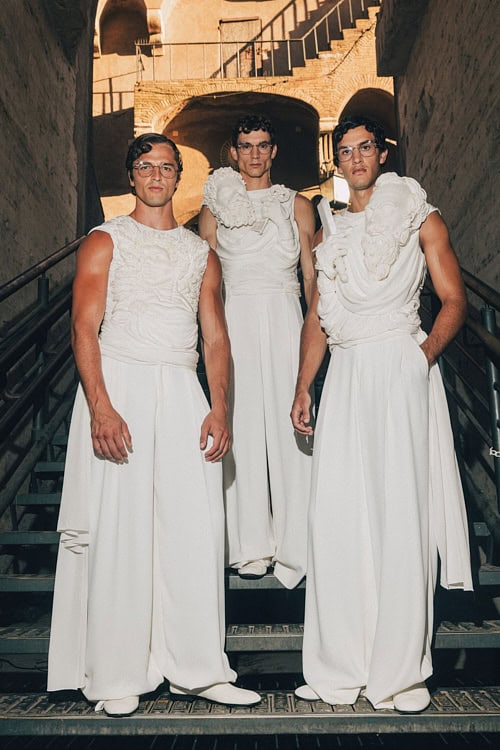 Three models in sculptural white sleeveless gowns on stairs, blending classical and modern – Dolce & Gabbana Alta Sartoria 2025.