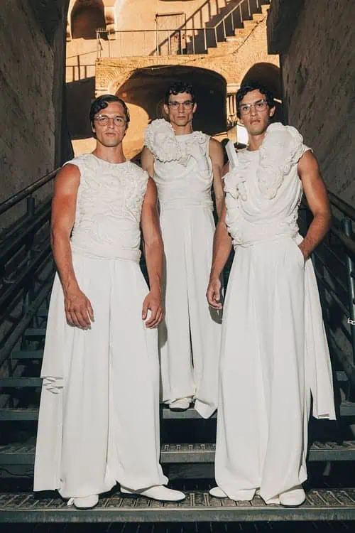 Three models in sculptural white sleeveless gowns on stairs, blending classical and modern – Dolce & Gabbana Alta Sartoria 2025.