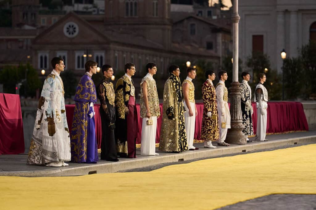 Lineup of male models in embroidered ceremonial looks on a yellow runway in front of Roman architecture – Dolce & Gabbana Alta Sartoria 2025.