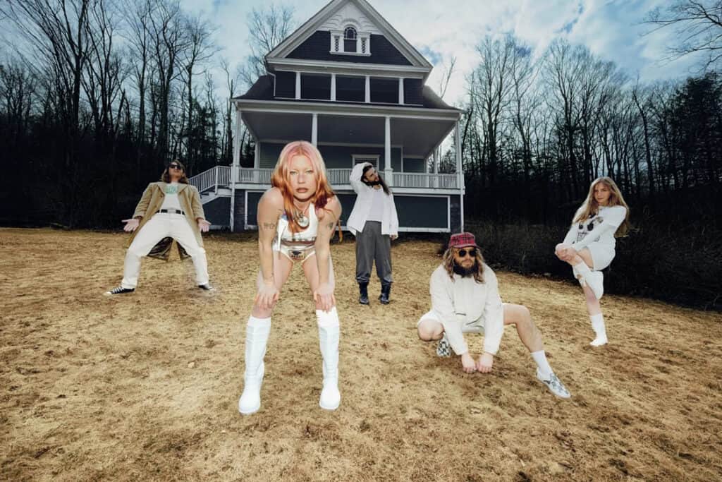 The band Wet Leg poses dramatically on a dried-out lawn in front of a large Victorian house, blending retro fashion with surreal, kinetic energy.