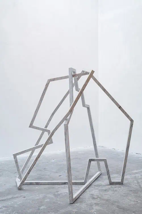 Geometric marble sculpture composed of intersecting angular frames in a minimalist gallery.