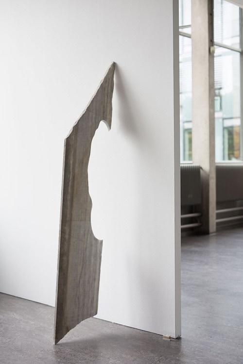 Thin marble slab with a carved organic contour hanging from the edge of a white gallery wall.