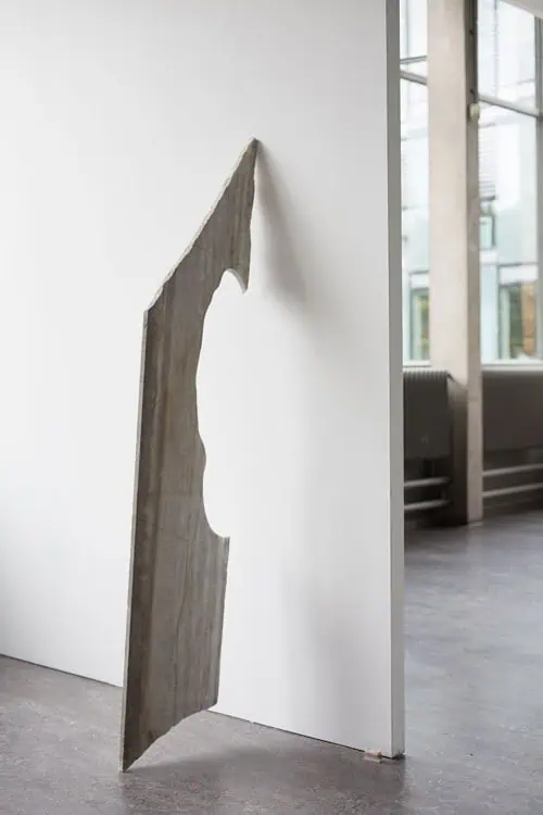 Thin marble slab with a carved organic contour hanging from the edge of a white gallery wall.