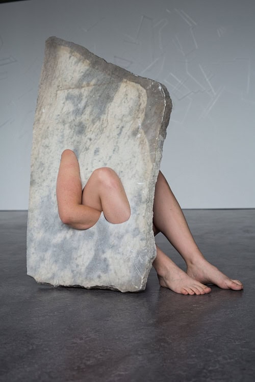 Large marble block with a limb extending through a carved cut-out shape, positioned on the floor.