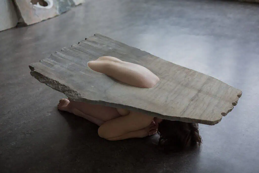 Human body curled beneath a heavy marble slab with a single limb protruding through a carved opening.