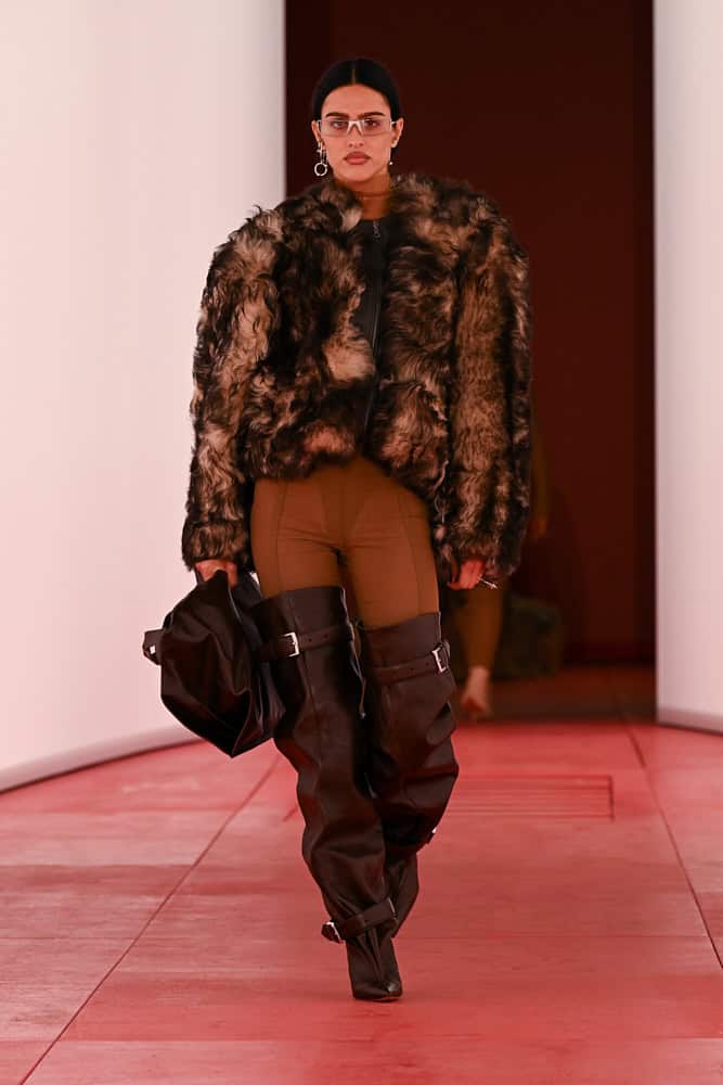 Model walking H&M runway in faux-fur jacket and thigh-high leather boots.