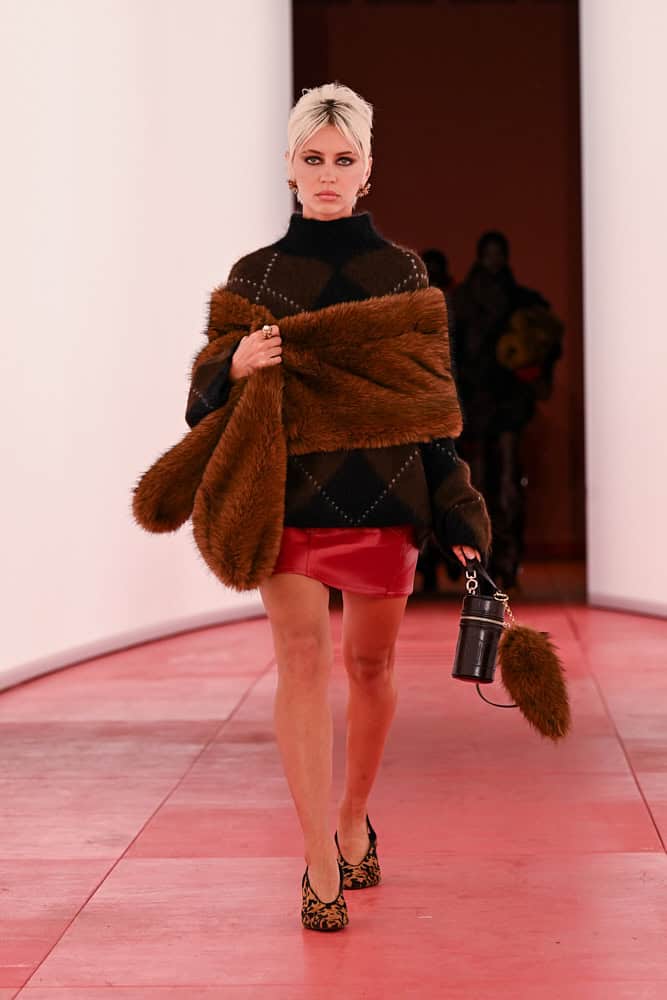 Model on H&M runway wearing argyle sweater, red mini skirt, and faux-fur scarf.
