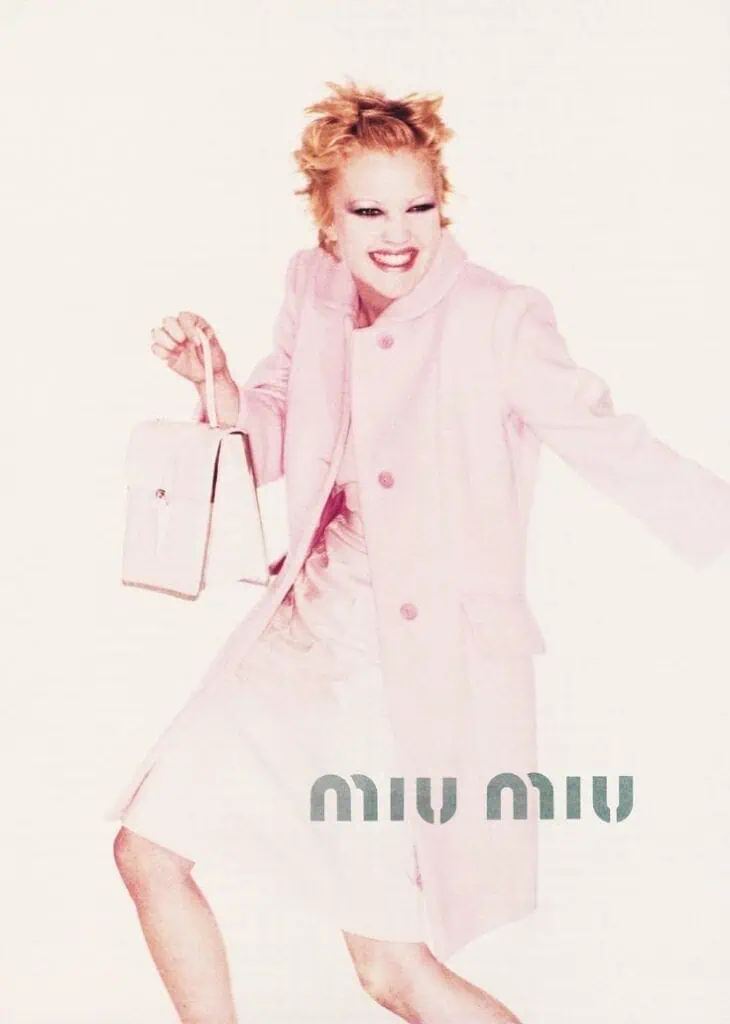 Fashion photograph from the Miu Miu Fall/Winter 1995 campaign featuring a model in a pale pink coat holding a handbag and smiling brightly.