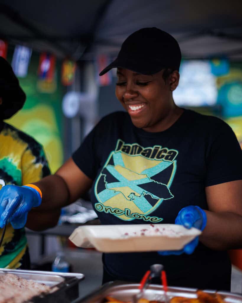 Food vendor in a Jamaica ‘One Love’ shirt serving Caribbean dishes at Notting Hill Carnival.