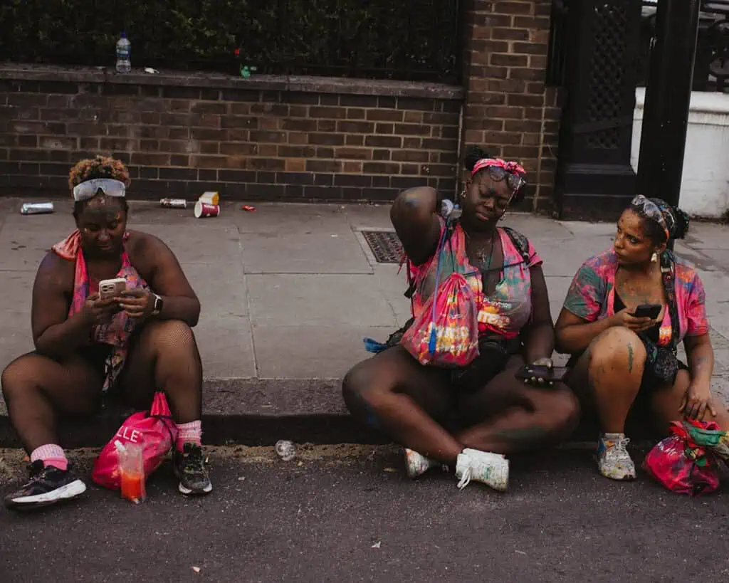Carnival-goers resting on the curb covered in paint after J’Ouvert at Notting Hill Carnival.