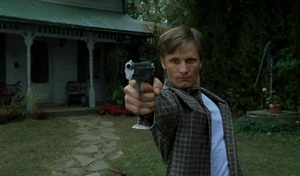 Still from A History of Violence (2005) showing a man aiming a gun in front of a suburban house, capturing intensity and confrontation.