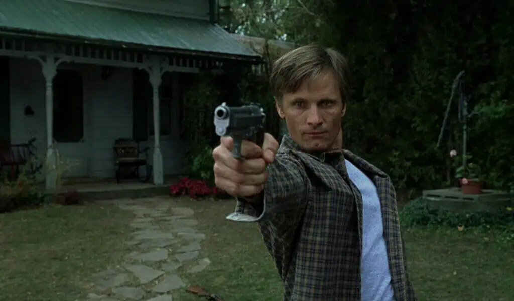 Still from A History of Violence (2005) showing a man aiming a gun in front of a suburban house, capturing intensity and confrontation.