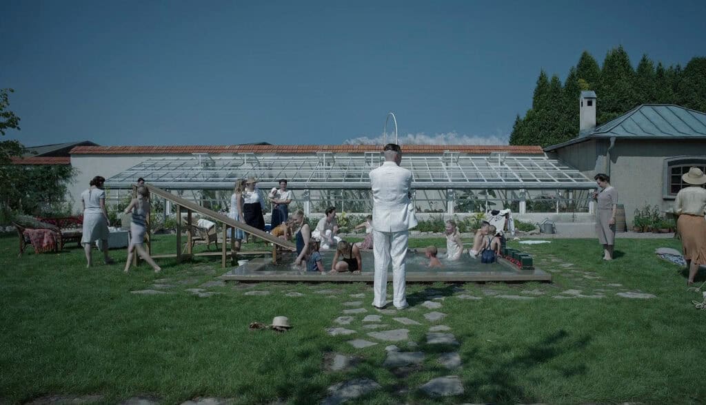 Scene from The Zone of Interest (2023) showing a family enjoying a sunny afternoon by a small pool in a garden beside a greenhouse, contrasting domestic calm with unseen horror.