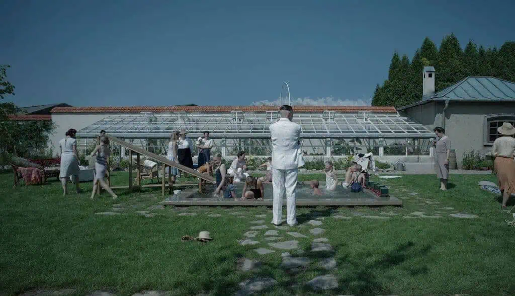 Scene from The Zone of Interest (2023) showing a family enjoying a sunny afternoon by a small pool in a garden beside a greenhouse, contrasting domestic calm with unseen horror.