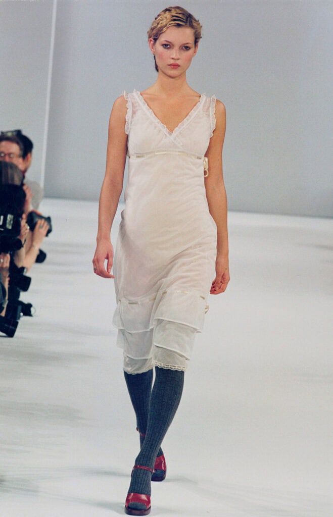 Runway image from Miu Miu Fall 1996 showing a model in a delicate white slip dress paired with grey knee socks and red heels.
