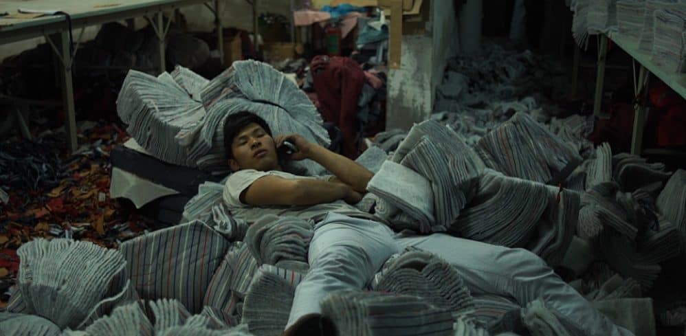 Young man resting among fabric piles in garment workshop, Wang Bing’s Youth documentary.