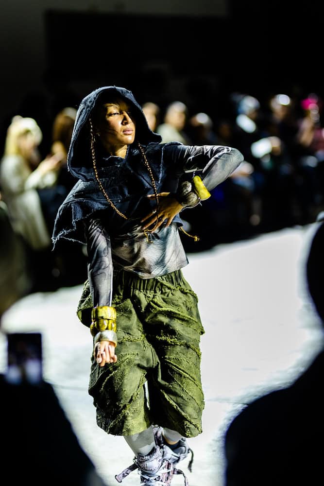 Model in layered hooded look with sculptural olive-green trousers on the runway, photographed by Hannah Khatib for Yaku Stapleton’s “A Ground to Stand On.”