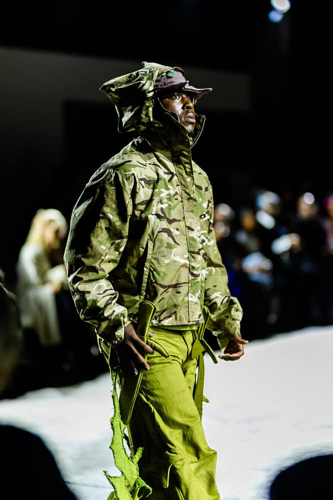 Model in camouflage jacket and neon-green pants on the catwalk, photographed by Hannah Khatib during Yaku Stapleton’s “A Ground to Stand On.”