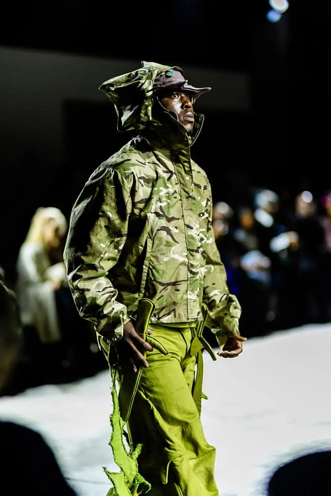 Model in camouflage jacket and neon-green pants on the catwalk, photographed by Hannah Khatib during Yaku Stapleton’s “A Ground to Stand On.”