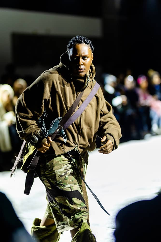 Model in oversized brown hoodie and camouflage trousers from Yaku Stapleton’s “A Ground to Stand On,” photo by Hannah Khatib.