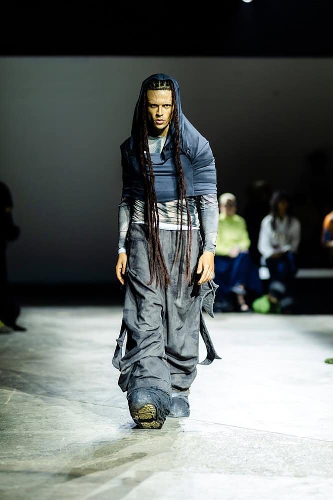 Model in layered navy garments with long braids walking the runway, from Yaku Stapleton’s “A Ground to Stand On,” shot by Hannah Khatib.
