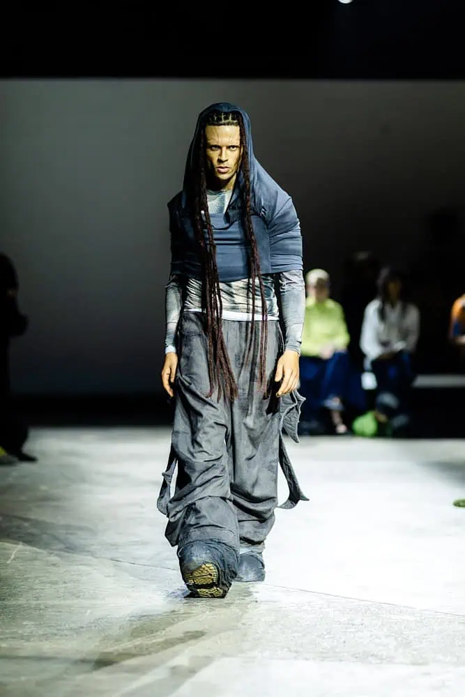 Model in layered navy garments with long braids walking the runway, from Yaku Stapleton’s “A Ground to Stand On,” shot by Hannah Khatib.