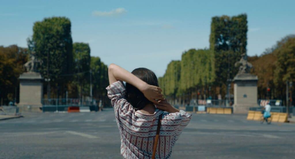 A scene from “Paul and Paulette Take a Bath,” showing Paulette standing in the middle of a Paris boulevard, hands behind her head.