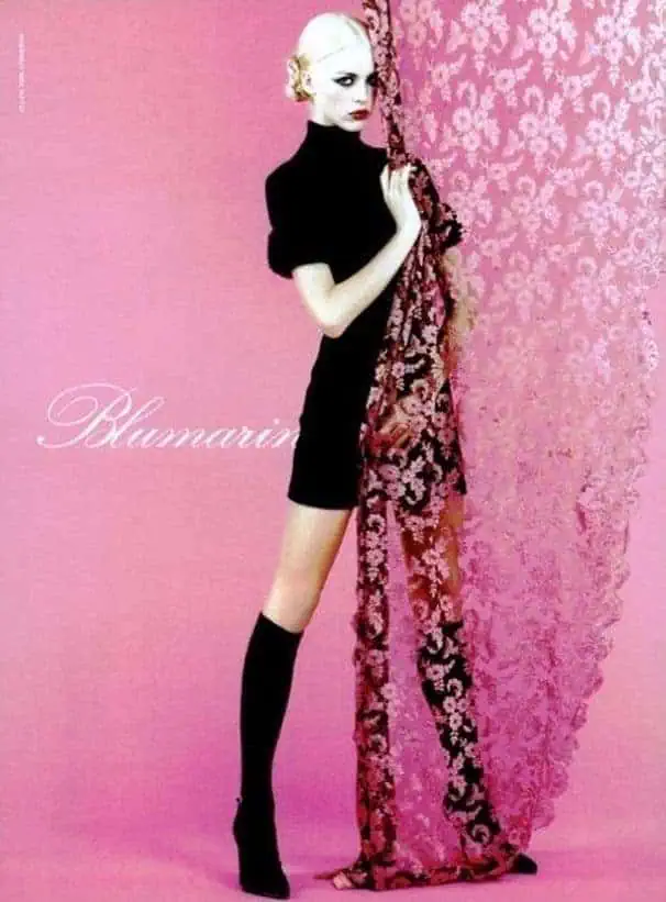 Editorial image from a Blumarine fashion campaign showing a model in a black dress and knee-high socks against a pink floral lace backdrop.