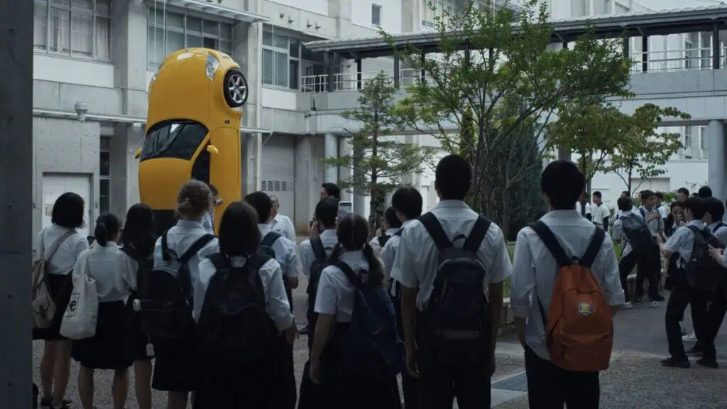 Students gather around a yellow car stood vertically on campus in Happyend.