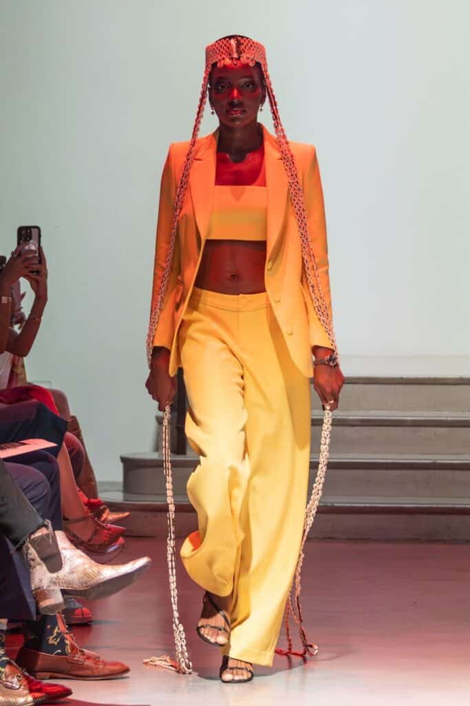A model walks the runway in a bright yellow suit from emerging designers, featuring a crop top, wide-leg pants, and open-toe sandals. She wears a beaded headpiece with long braids sweeping the floor as audience members watch on either side.