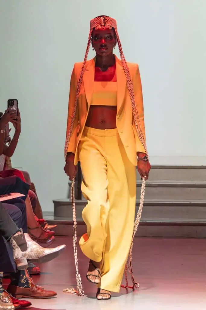 A model walks the runway in a bright yellow suit from emerging designers, featuring a crop top, wide-leg pants, and open-toe sandals. She wears a beaded headpiece with long braids sweeping the floor as audience members watch on either side.