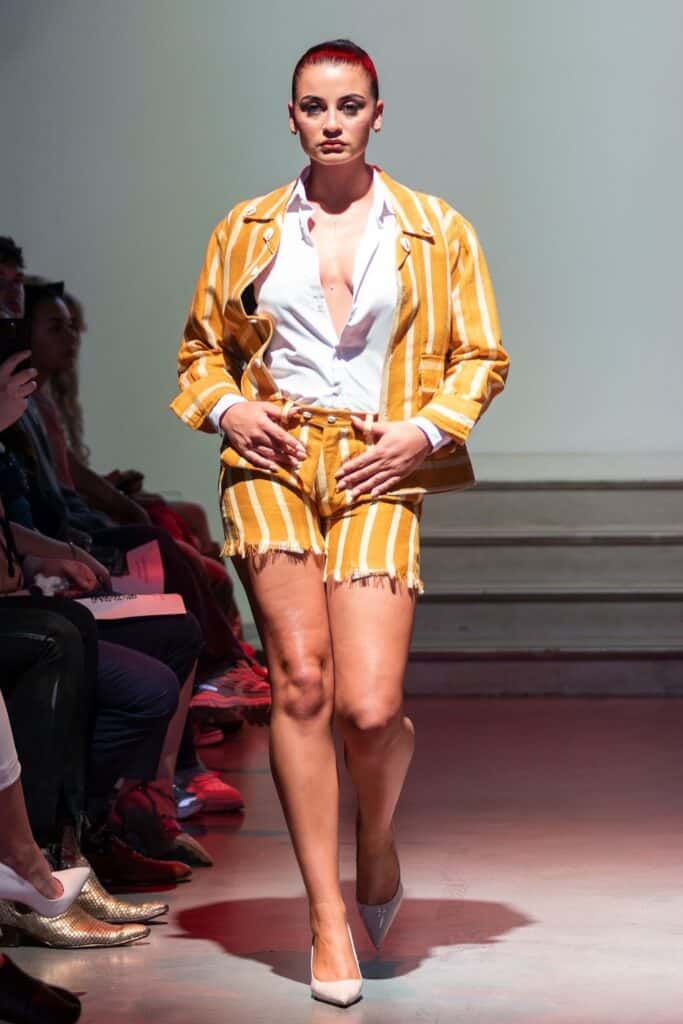 A model walks a runway for emerging designers, wearing a yellow and white striped blazer with matching frayed shorts and a white button-down shirt, partially unbuttoned. She has slicked-back red hair and white heels as audience members watch from both sides.