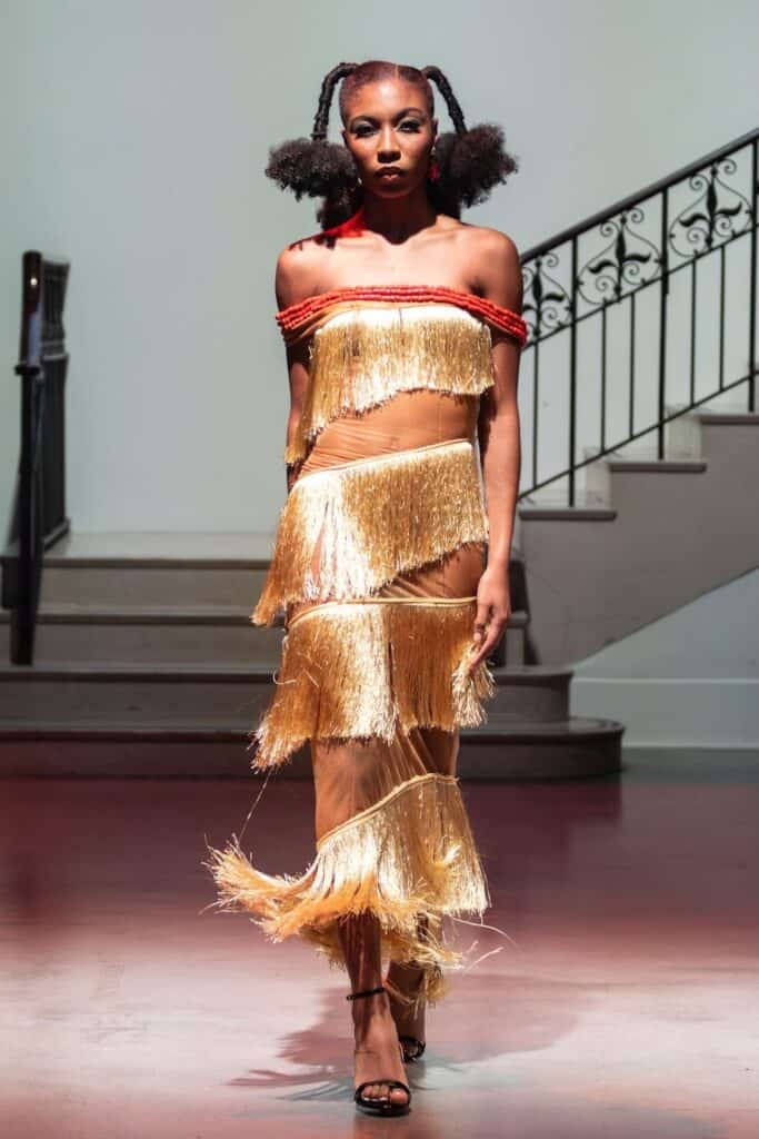 A model walks on a pink runway wearing a gold fringe two-piece from emerging designers, accented with red off-shoulder flair. She has two afro puffs and stands before a staircase with an ornate railing.