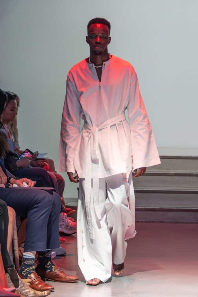 A male model walks a runway for emerging designers, wearing a loose-fitting white robe with wide sleeves, a waist tie, and flowing white pants. He is barefoot, and the audience is seated on either side of the catwalk.