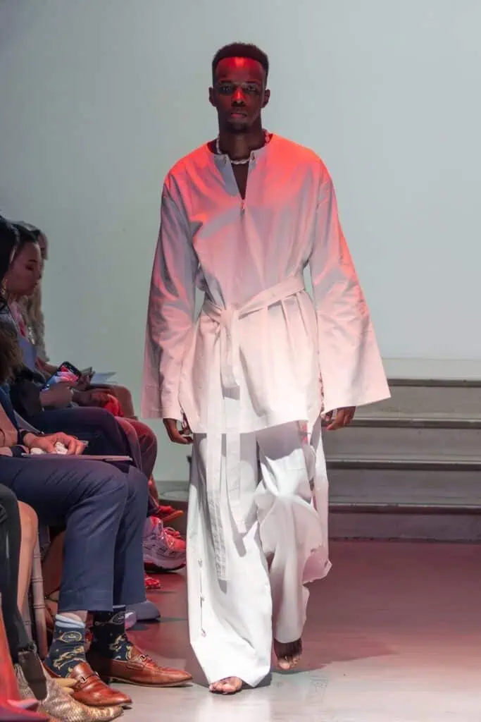 A male model walks a runway for emerging designers, wearing a loose-fitting white robe with wide sleeves, a waist tie, and flowing white pants. He is barefoot, and the audience is seated on either side of the catwalk.