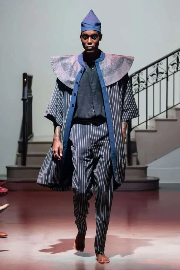 A barefoot model walks a runway for emerging designers, wearing a pinstriped outfit with wide-legged pants, a short-sleeved cape with structured shoulders, and a matching pointed hat, set in front of a staircase.