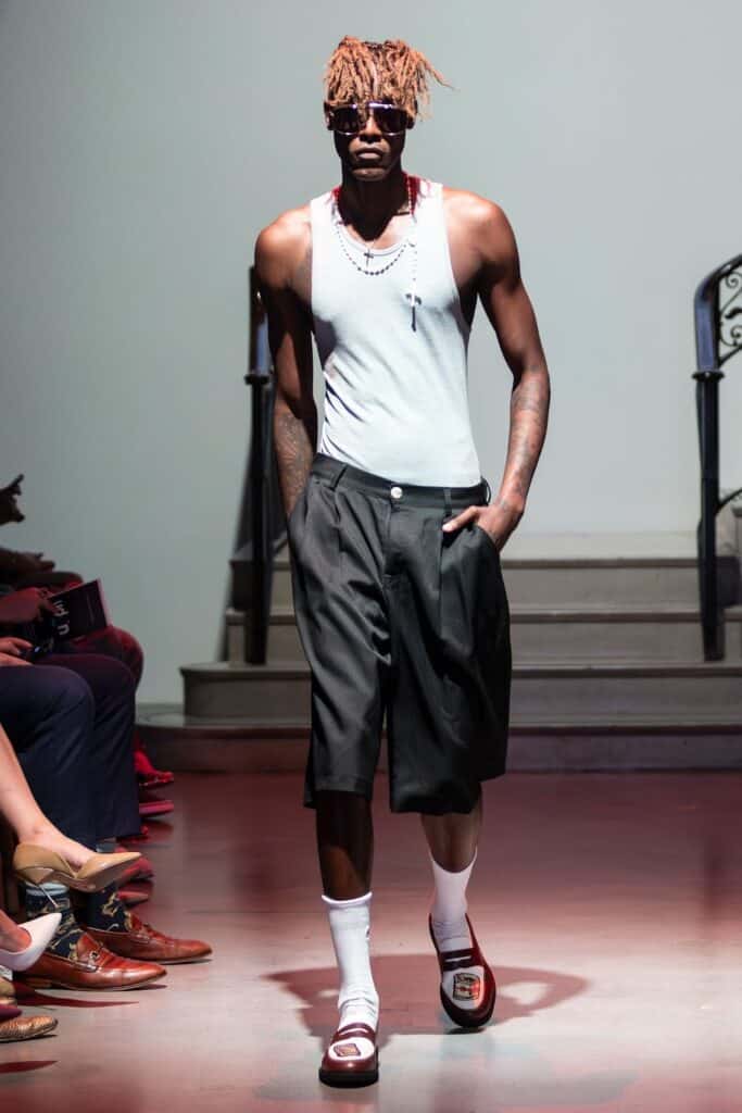 A male model with sunglasses and necklaces walks a runway in a look from emerging designers: white tank top, loose black knee-length shorts, long white socks, and red-and-white slide sandals. Stairs and seated spectators are visible in the background.