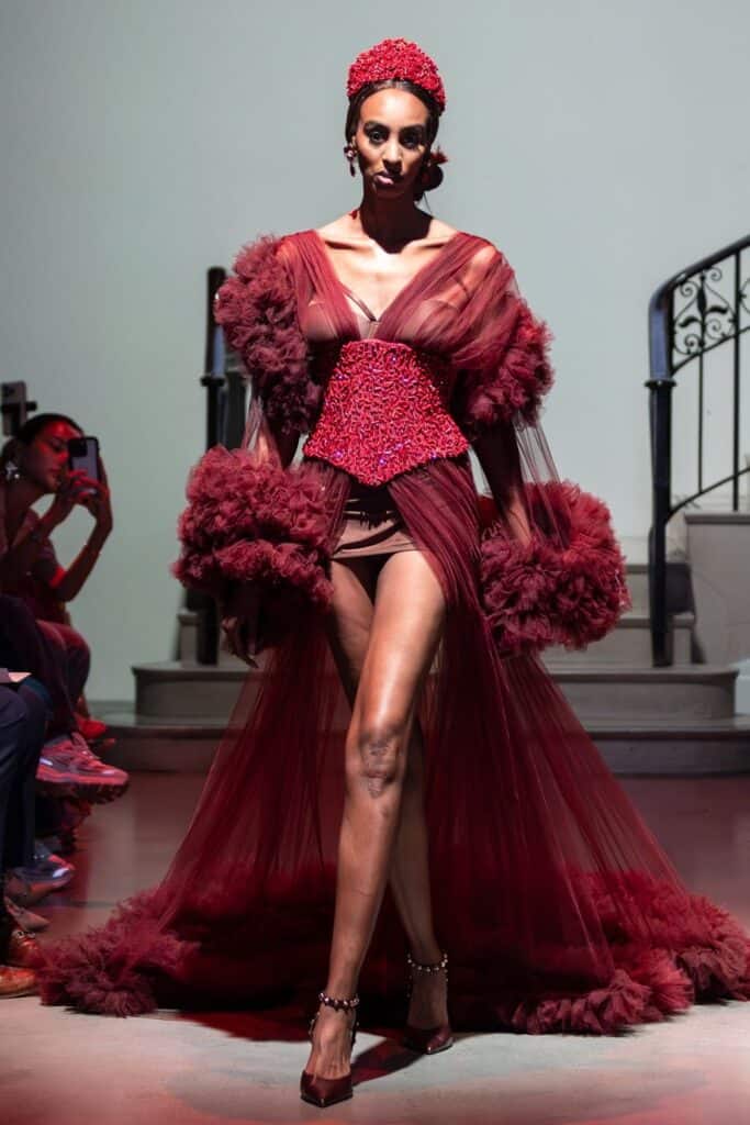A model walks a runway in a dramatic deep red sheer gown with voluminous tulle sleeves and skirt, a beaded corset, and jeweled headpiece—showcasing the artistry of emerging designers against a striking staircase background.