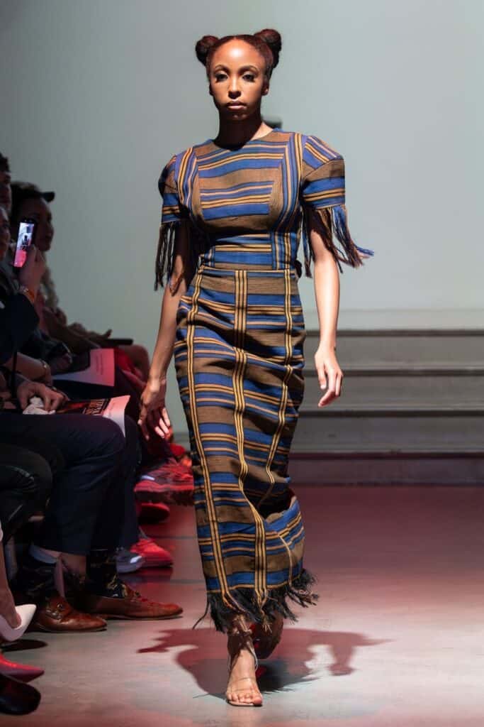 A model walks on a runway for emerging designers, wearing a fitted, ankle-length dress with blue, gold, and brown stripes, fringe details on the sleeves and hem, and her hair styled in two high buns. Audience members are seated on either side.