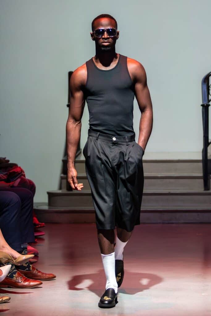 A male model wearing sunglasses, a black tank top, knee-length black pleated shorts, white socks, and black loafers walks on a runway with one hand in his pocket, showcasing the bold vision of emerging designers.