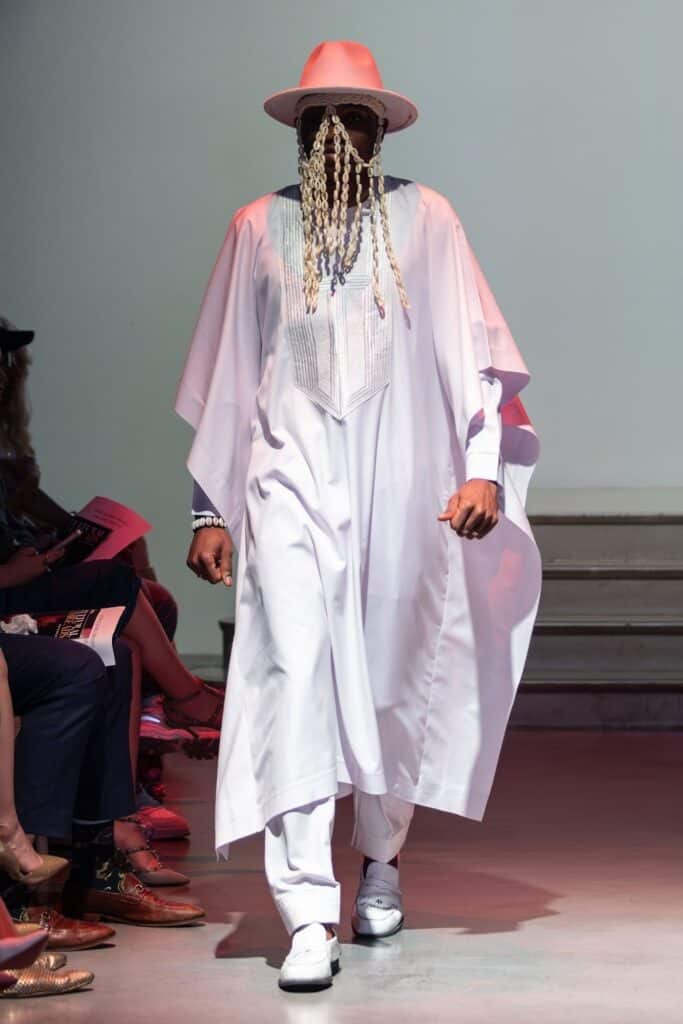 A model for emerging designers walks the runway in a flowing white robe, white pants, white shoes, a wide-brimmed hat, sunglasses, and long beaded braids covering the face. The audience is visible seated on either side.