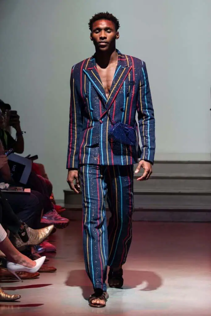 A male model walks down a runway wearing a dark suit with colorful vertical stripes, open at the chest, paired with matching pants and sandals—a bold look from emerging designers. Audience members on either side watch the fashion show intently.