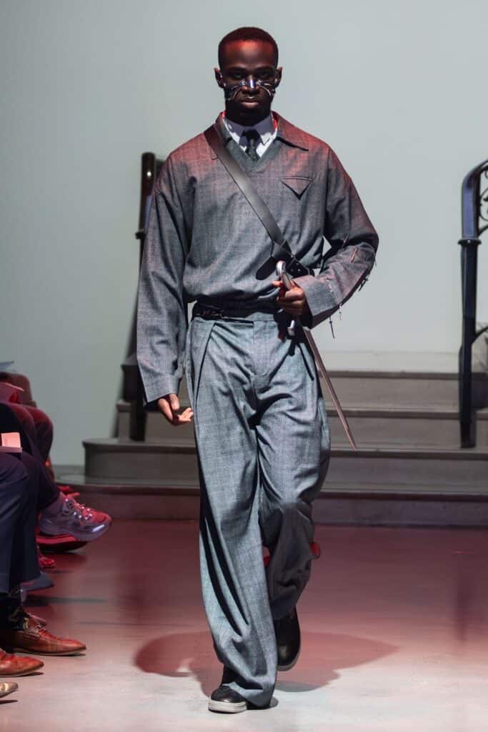 A model wearing a loose-fitting gray suit by emerging designers, with a collared shirt, tie, and crossbody strap, walks down the runway. The look features wide-leg pants, long sleeves, and a striking long silver cane.