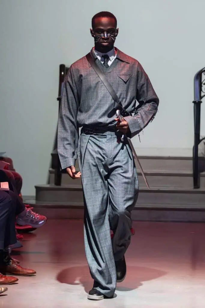 A model wearing a loose-fitting gray suit by emerging designers, with a collared shirt, tie, and crossbody strap, walks down the runway. The look features wide-leg pants, long sleeves, and a striking long silver cane.