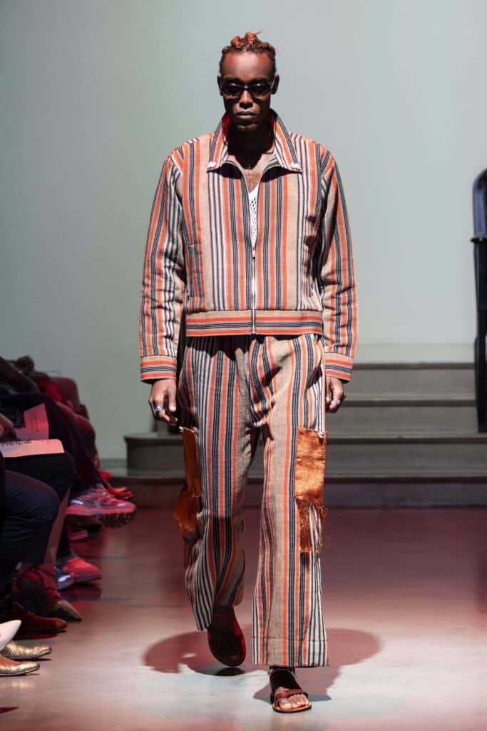 A model walks a runway wearing a striped, multicolored jacket and matching pants with fringe accents, showcasing bold orange, beige, and black stripes. This striking look from emerging designers is completed with sunglasses and sandals.
