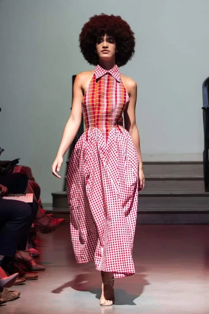 A model with curly hair walks a runway in a sleeveless dress by emerging designers, featuring a fitted, multicolored plaid top and a full red-and-white gingham skirt. The background is minimal with steps and soft lighting.