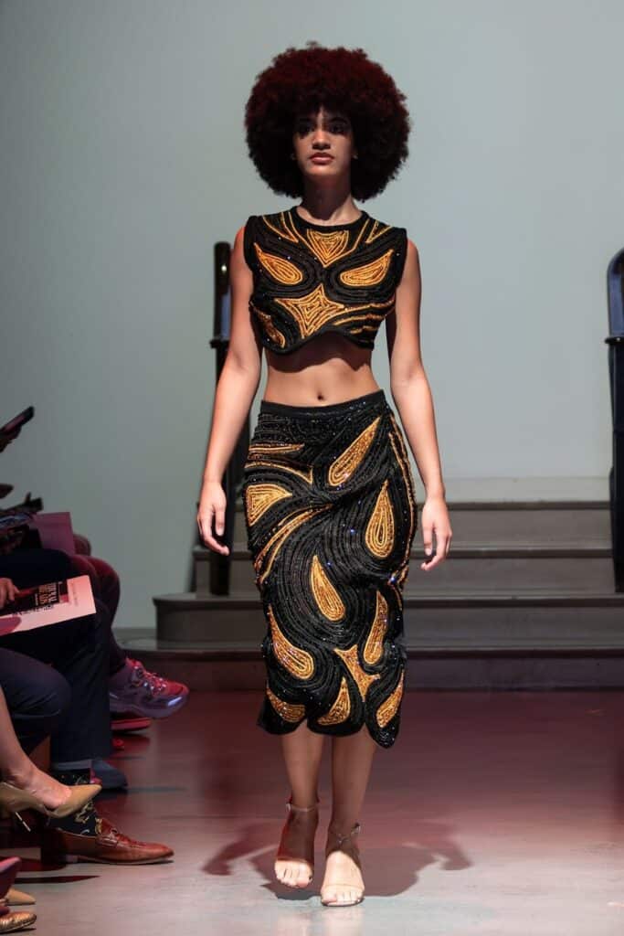 A model with an afro hairstyle walks the runway in a striking two-piece outfit by emerging designers, featuring black and gold swirling, paisley-like patterns. The sleeveless crop top and knee-length skirt impress the audience seated along the sides.