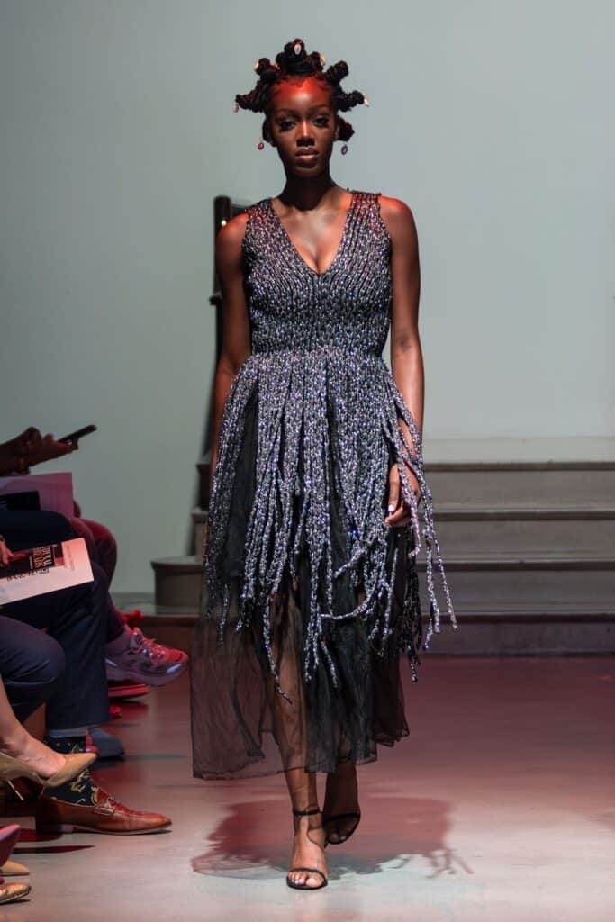 A model with textured hair in bantu knots walks a runway wearing a sleeveless, deep V-neck dress by emerging designers, featuring a beaded, fringed skirt overlaying a sheer black layer. The setting has a modern, minimalistic backdrop with steps.