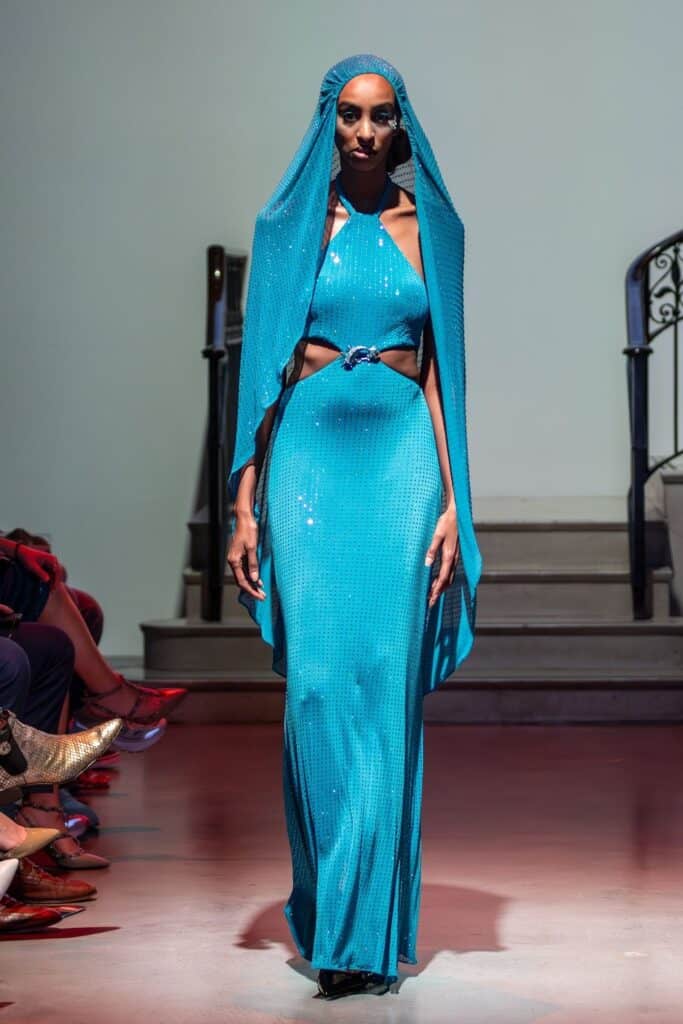 A model walks a runway for emerging designers, wearing a shimmering turquoise gown with a belted waist, cut-out sides, and an attached matching hood, under bright lighting. The audience is seated along the sides with a staircase in the background.